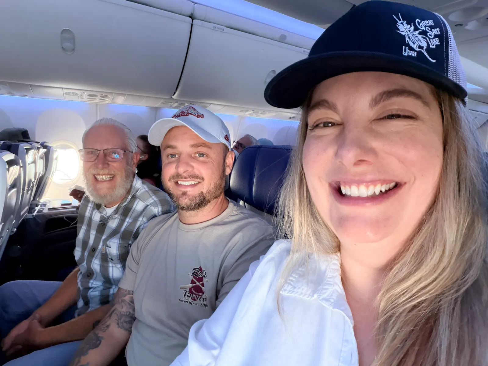 Selfie of three smiling passengers in an airplane row, one wearing a Ray's Tavern Green River shirt