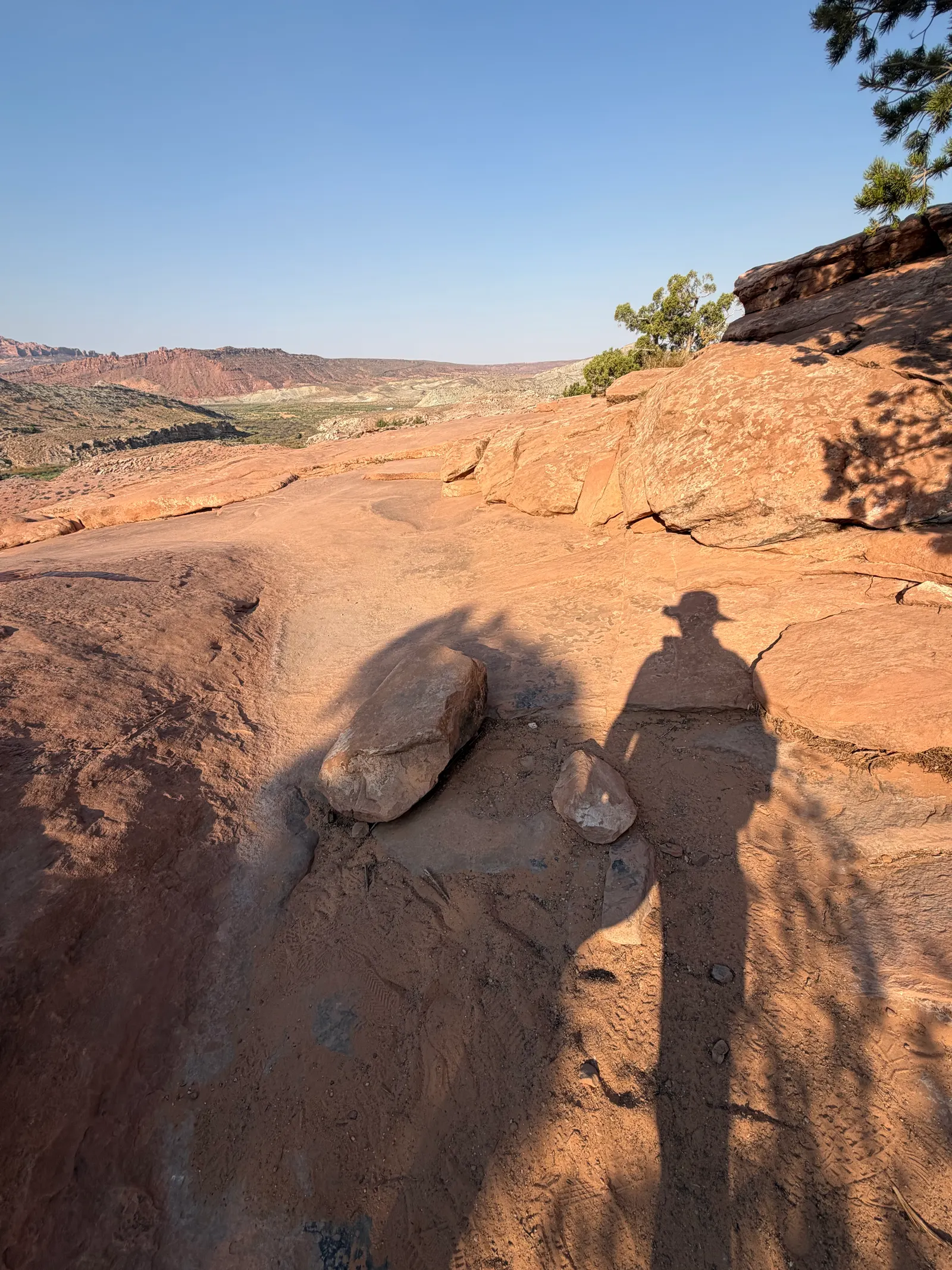 Long morning shadow of a man in a hat cast across the slickrock, valley hazy below